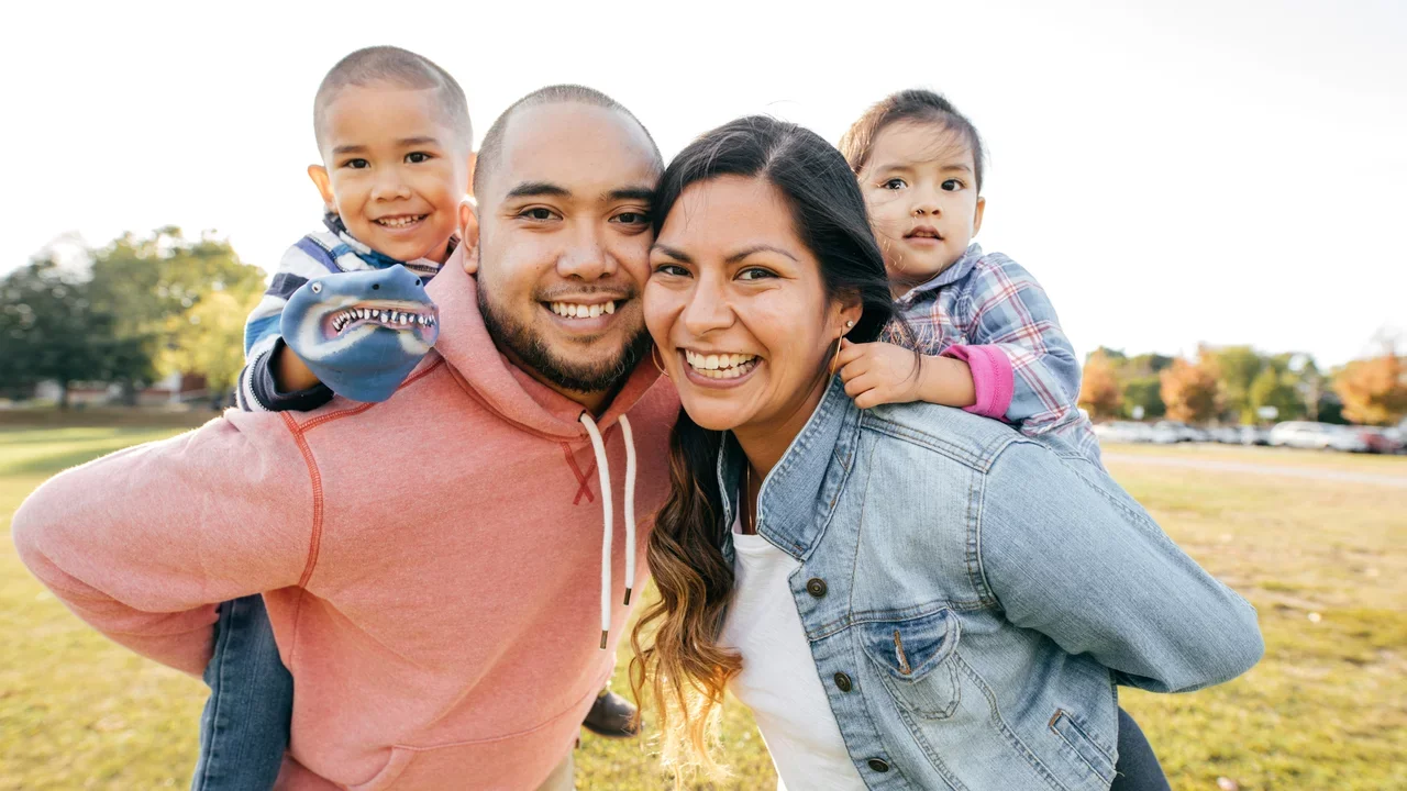 Photograph of a family of four, smiling, while the kids are on the backs of the parents.
