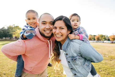 Photograph of a family of four, smiling, while the kids are on the backs of the parents.