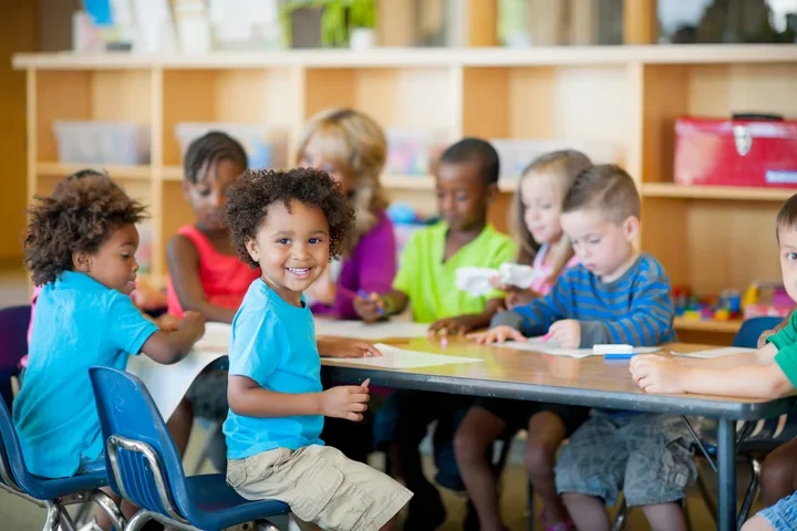 Photograph of a group of children smiling at the camera while sitting in a classroom.