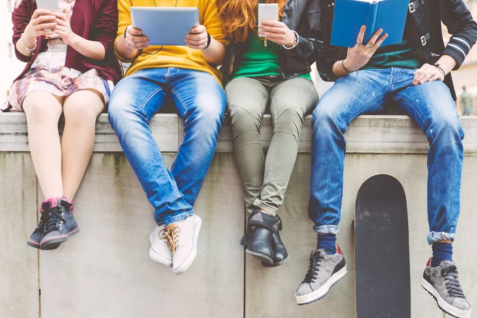 Photograph of four young kids sitting on a wall, all reading books or using electronic devices. Heads of children are omitted from the photo.