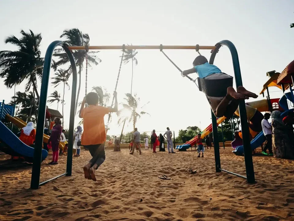 Photograph of two kids swinging on swings with their backs to the camera in the foreground. Playgound in the background and sun shining with subtle lens flare overlaid on photo.